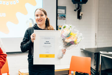 A smiling person is holding an honorary diploma and a bouquet of flowers.