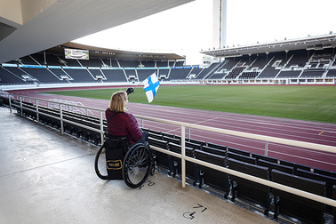 Nainen istuu pyörätuolissa Stadionin katsomossa ja heiluttaa Suomen lippua.