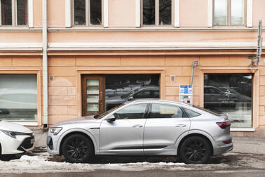 Harmaa henkilöauto pysäköitynä kadulle, taustalla oranssi kerrostalo.