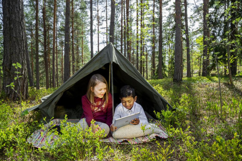 Kaksi lasta lukee e-kirjaa teltassa metsän keskellä.