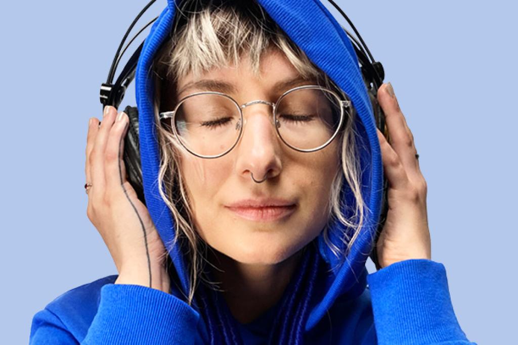 Sinisiin vaatteisiin pukeutunut nainen kuuntelee musiikkia silmät kiinni kuulokkeiden kautta. 