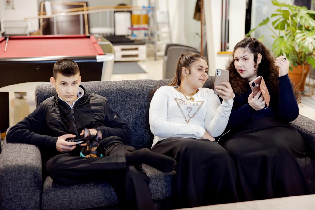 kolme romaninuorta istuvat sohvalla. poika pelaa ja kaksi tyttöä keskustelee ja katselee puhelimiaan.