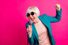 Aurinkolasipäinen nainen laulaa mikrofoniin pinkin seinän edessä.