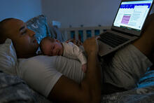 Aikuinen lepää sängyssä sylissään vauva ja kannettava tietokone. 