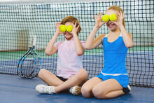 Kaksi lasta hassuttelee tenniskentällä pitämällä palloja silmiensä edessä.